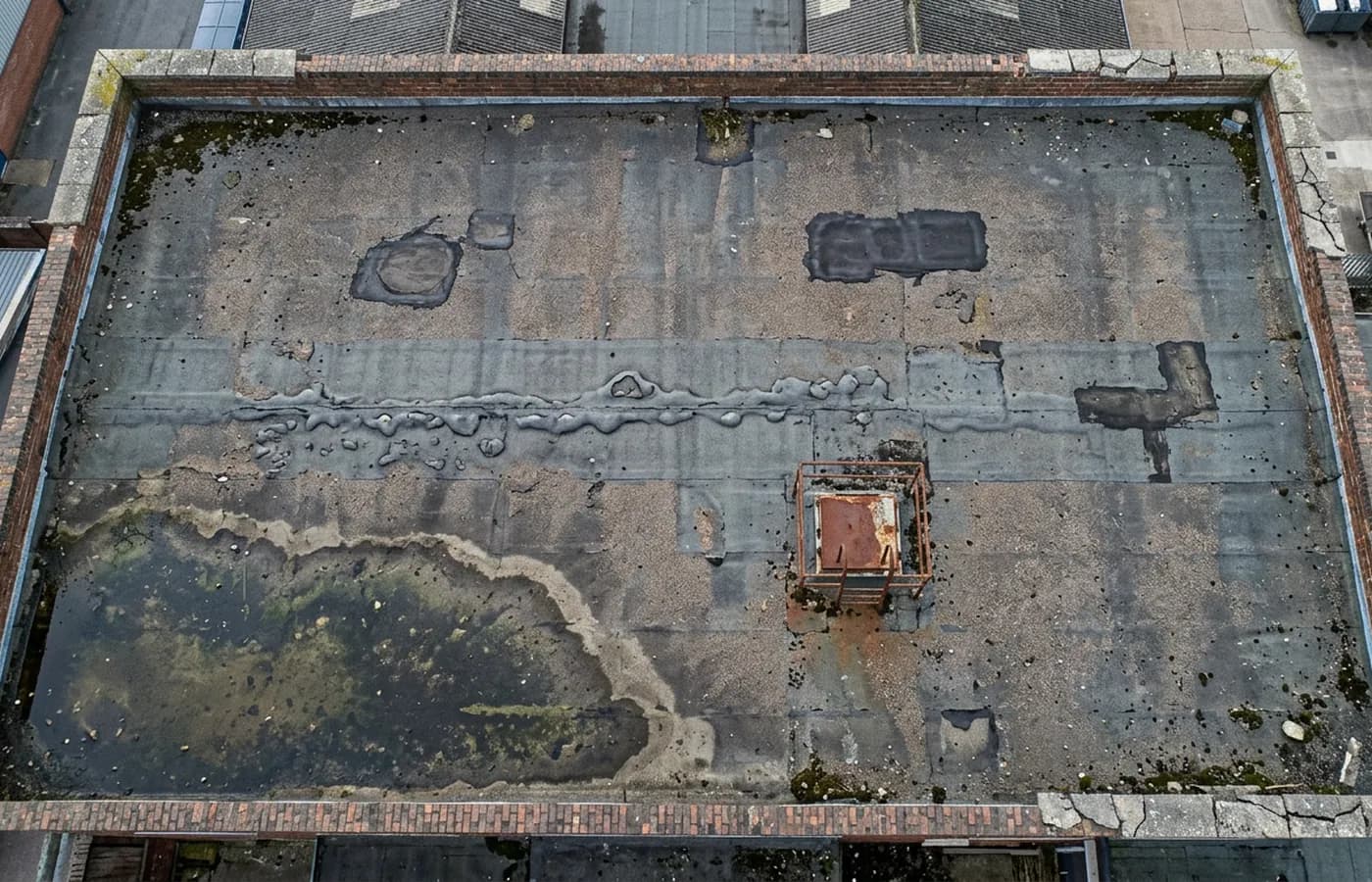 An ageing flat commercial roof photographed from directly above before refurbishment, with patch repairs, blistering and a ponding area